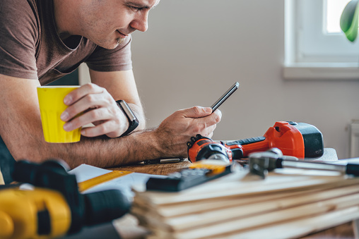 Man using smart phone and holding yellow cup of coffee in front of table with tools at home