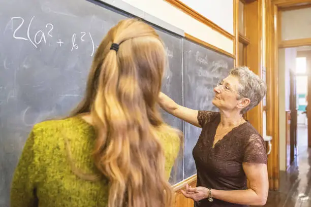 Senior female teacher at chalkboard explaining math formula to female teenage student. Personal Perspective, Selective focus, small DOF. Natural interior light.