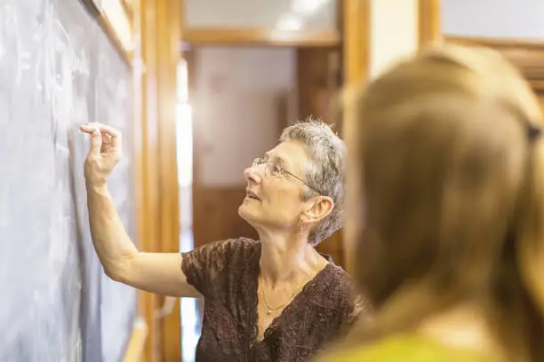 Senior female teacher at chalkboard explaining math formula to female teenage student. Personal Perspective, Selective focus, small DOF. Natural interior light.