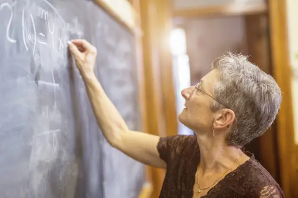 Concentrated senior female mathemathics professor writing a formula to the chalkboard.. Personal Perspective, Selective focus, small DOF. Natural interior light.