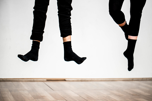 Close up image of two pairs of legs, wearing black pants and black socks beyond the parquet. Both persons are in jump and one has flat feet and the other has crossed with points. White wall in the background. Shot made in Ljubljana, Slovenia, Europe.