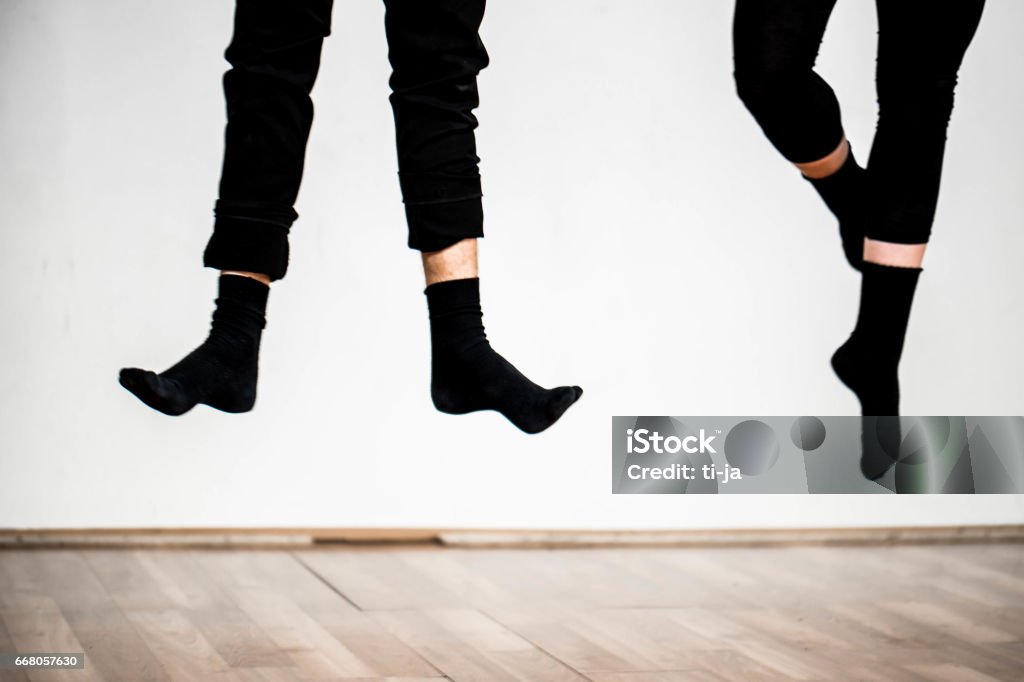 Happy feet Close up image of two pairs of legs, wearing black pants and black socks beyond the parquet. Both persons are in jump and one has flat feet and the other has crossed with points. White wall in the background. Shot made in Ljubljana, Slovenia, Europe. Dancing Stock Photo