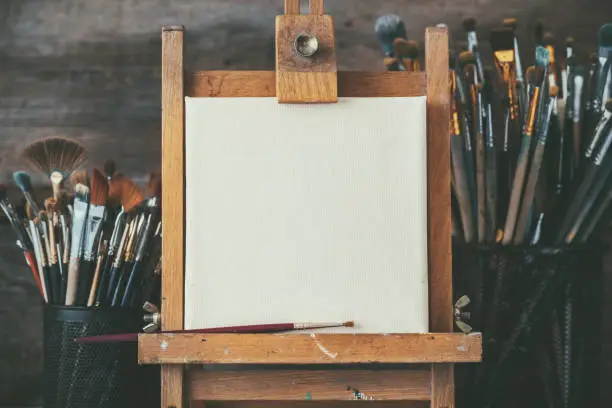 Photo of Artistic equipment in a artist studio: empty artist canvas on wooden easel and paint brushes Retro toned photo.