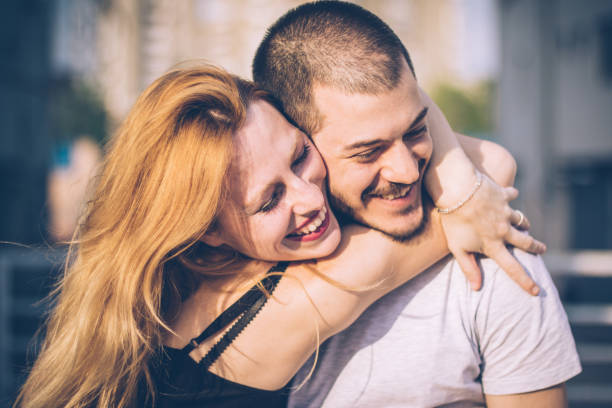 A smiling woman hugs a man from behind in a park, their expressions reflecting happiness and contentment as they stand amidst greenery, bathed in the soft glow of the evening sun.