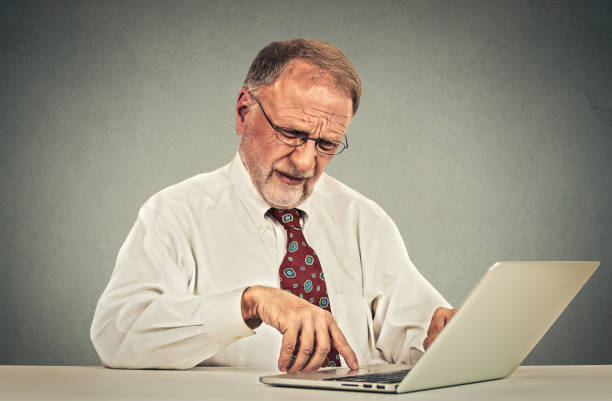 istock Confused looking elderly mature man with glasses sitting at table working typing on laptop computer 668090560