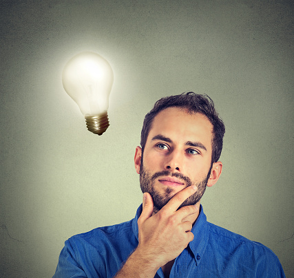 Closeup portrait man thinks looking up at bright light bulb isolated on gray wall background. Idea and people concept. Human face expression