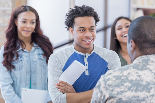 Attentive young African American man talks with an armed forces recruitment officer at a recruitment event. The young man is holding a brochure.