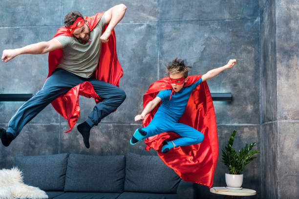 father and son in superhero costumes jumping on sofa at home father and son in superhero costumes jumping on sofa at home heroes photos stock pictures, royalty-free photos & images