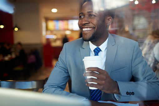 Portrait of cheerful African âAmerican businessman drinking coffee from disposable cup and looking away smiling  in coffee shop, shot behind glass window