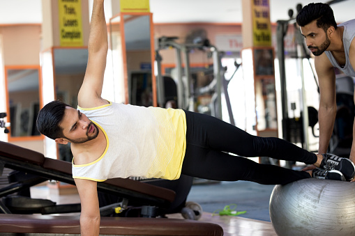 Trainer with young man in side plank pose on Swiss ball at gym