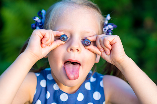 Smiling girl holding blueberries in front of her face - showing her tongue - covering eyes with berries