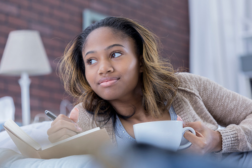 Pretty young woman looks to the side while she daydreams. She is writing in a journal. She is holding a coffee cup.