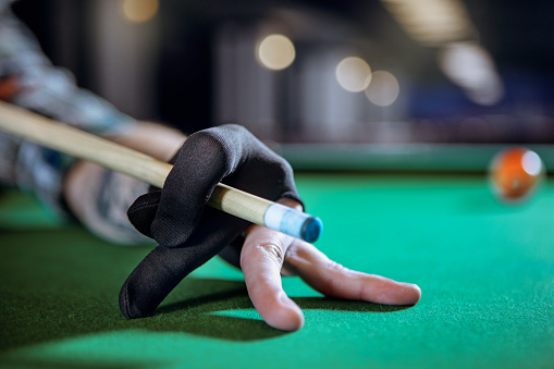 Player's hand is preparing to hit the ball. On the photo there are a green billiard table, cue and player's hand in black sport glove. On the tip of the cue is a blue chalk.