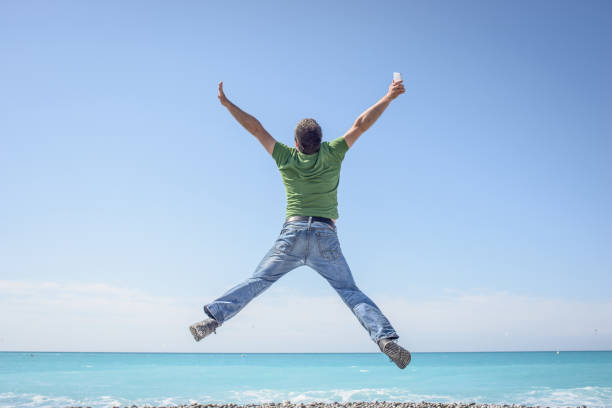Man so happy with his mobile / cell phone and network that he has to express his contentment by jumping in the air. It's a beautiful sunny summer's day on the pebble beach in Nice, France. Lots of blue sky, great copy space.