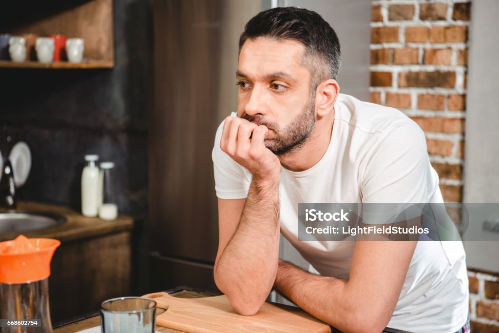 Thoughtful man in kitchen Thoughtful man leaning on kitchen table and looking away Introspection Stock Photo