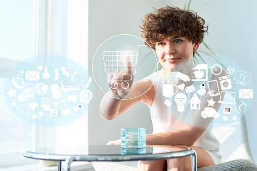 Young woman sitting at home and doing online shopping on transparent touchscreen by pressing button with shopping cart image
