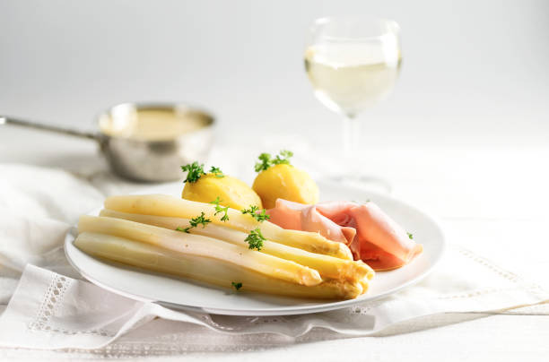white asparagus, potatoes, ham, sauce hollandaise and wine, bright background Juicy white asparagus dish with potatoes and ham on a white plate, sauce hollandaise and wine blurred in the bright background, copy space, selected focus, narrow depth of field hollandaise sauce stock pictures, royalty-free photos & images