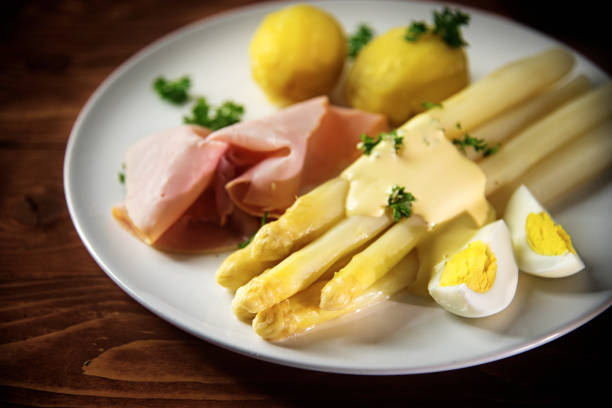 White asparagus dish with potatoes, ham, egg and sauce on a white plate, rustic reddish wooden table, selective focus, narrow depth of field