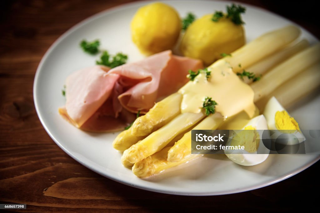 White asparagus  with potatoes, ham, egg and sauce, dark wood White asparagus dish with potatoes, ham, egg and sauce on a white plate, rustic reddish wooden table, selective focus, narrow depth of field Egg - Food Stock Photo