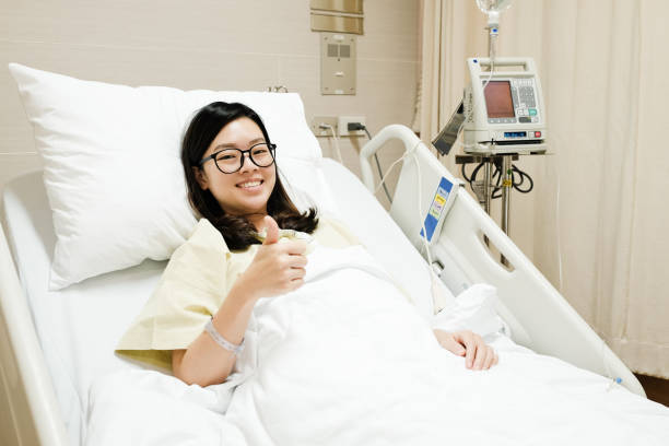 Asian patient girl thumbs up ok sign and smile while resting on hospital bed, positive medical concept stock photo