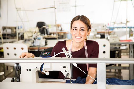 Portrait of a gorgeous young seamstress sitting in front of a sewing machine in a textile factory and smiling