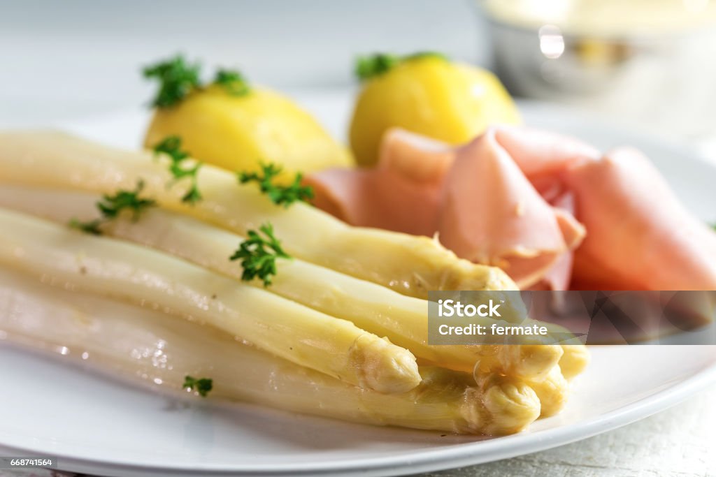 white asparagus, potatoes, ham, sauce hollandaise on a white plate Fresh white asparagus dish with potatoes and ham, light background, close-up, selective focus, narrow depth of fieldJuicy white asparagus dish with potatoes and ham on a white plate, sauce hollandaise and wine blurred in the bright background, copy space, selected focus, narrow depth of field Asparagus Stock Photo
