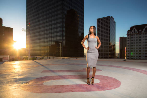 The sun is setting between skyscrapers and an elegant woman in a gray dress standing on a rooftop helipad.