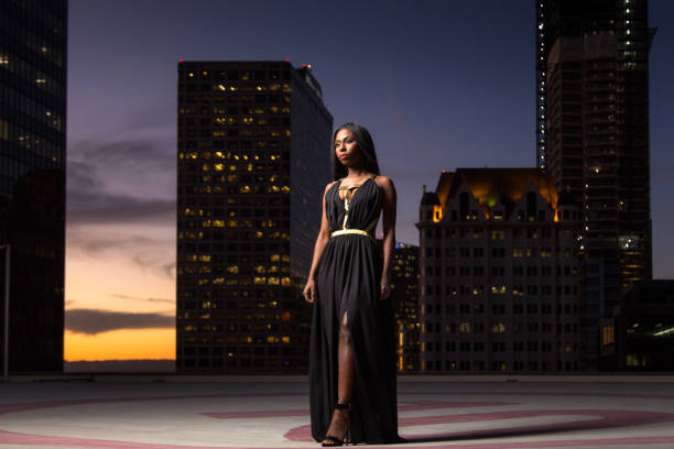 Portrait of an elegant African-American woman wearing a gray and gold Grecian style dress, standing on a skyscraper rooftop at dusk.