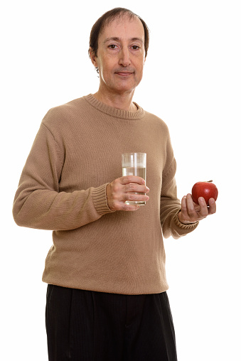 Studio shot of mature man standing while holding glass of water and red apple vertical shot