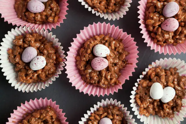 Close up of chocolate rice krispie cakes in pink and white cup cake cases with sugar coated eggs on top on a black slate background.