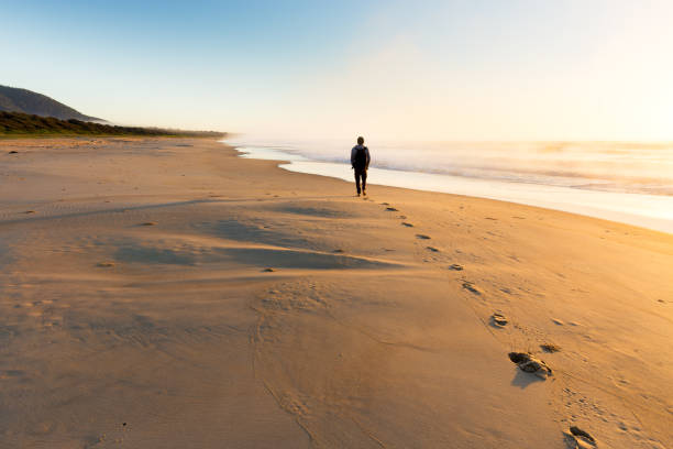 person zu fuß entlang einer misty strand bei sonnenaufgang - footprint sand sea beach stock-fotos und bilder