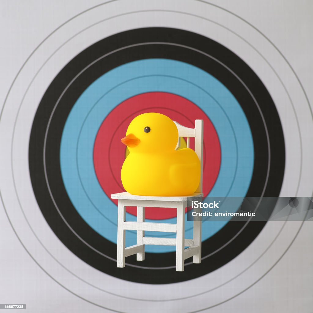 Sitting yellow rubber duck sitting on a chair in front of the bulls-eye of a sports target. A yellow rubber duck sitting on a chair in front of a sports target, concept image relating to a sitting duck, vulnerability, easy target etc. Duck - Bird Stock Photo