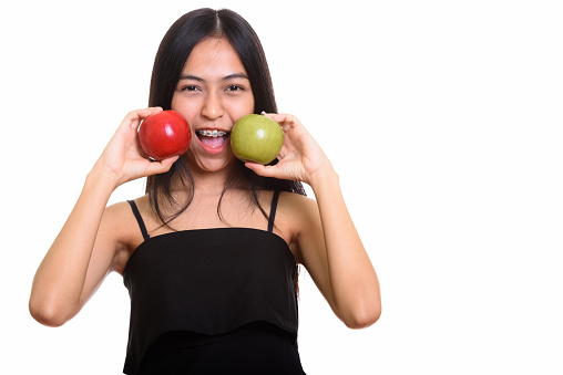 Studio shot of young happy Asian teenage girl smiling while putting green and red apple near the face horizontal shot