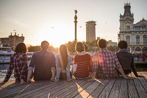 Rear view of males and females sitting in row on boardwalk. Men and women are wearing casuals at harbor. They are looking at city.