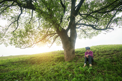 girl with VR headset sitting in grass field under big tree near sunset