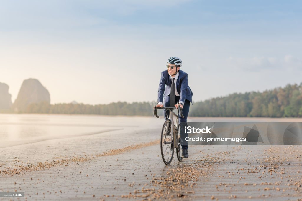 Giovane uomo d'affari asiatico in bicicletta rilassando la spiaggia al tramonto - Foto stock royalty-free di Abbigliamento da lavoro