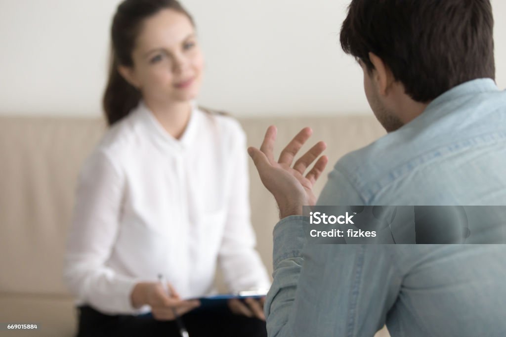 Female psychologist consulting male patient, job interview, business meeting Smiling female psychologist working with young man, business people talking, cheerful businesswoman or secretary in a white shirt holding clipboard and taking notes, applying for a job interview Mental Health Professional Stock Photo