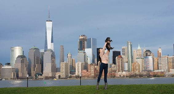 Young woman holding a mature black Pug dog in a pretty park with the New York City Skyline in the background.  Taken on a sunny spring day.