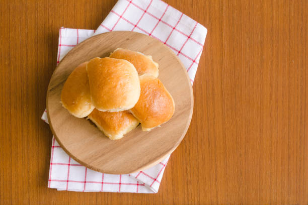 Fresh homemade baking delicious butter bun bread over wooden tray on red and white napkin on brown teakwood floor background with sunlight on the side for happy breakfast with copy space on the right Fresh homemade baking delicious butter bun bread over wooden tray on red and white napkin on brown teakwood floor background with sunlight on the side for happy breakfast with copy space on the right bun bread stock pictures, royalty-free photos & images