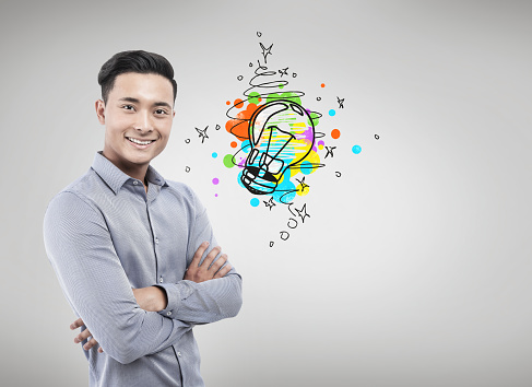 Portrait of a smiling Asian businessman and standing with crossed arms near a concrete wall with a small light bulb icon. Mock up