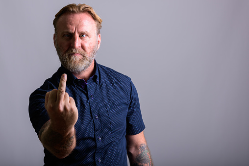 Studio shot of mature bearded man with hand tattoos showing his middle finger in gray background horizontal shot