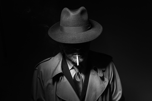 Man with fedora hat and trench coat smoking a cigarette in the dark, 1950s noir film character