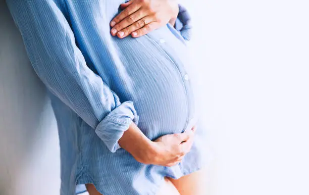 Pregnant woman holds hands on belly at home interiors. Pregnancy, parenthood, preparation and expectation concept. Close-up, copy space, indoors.