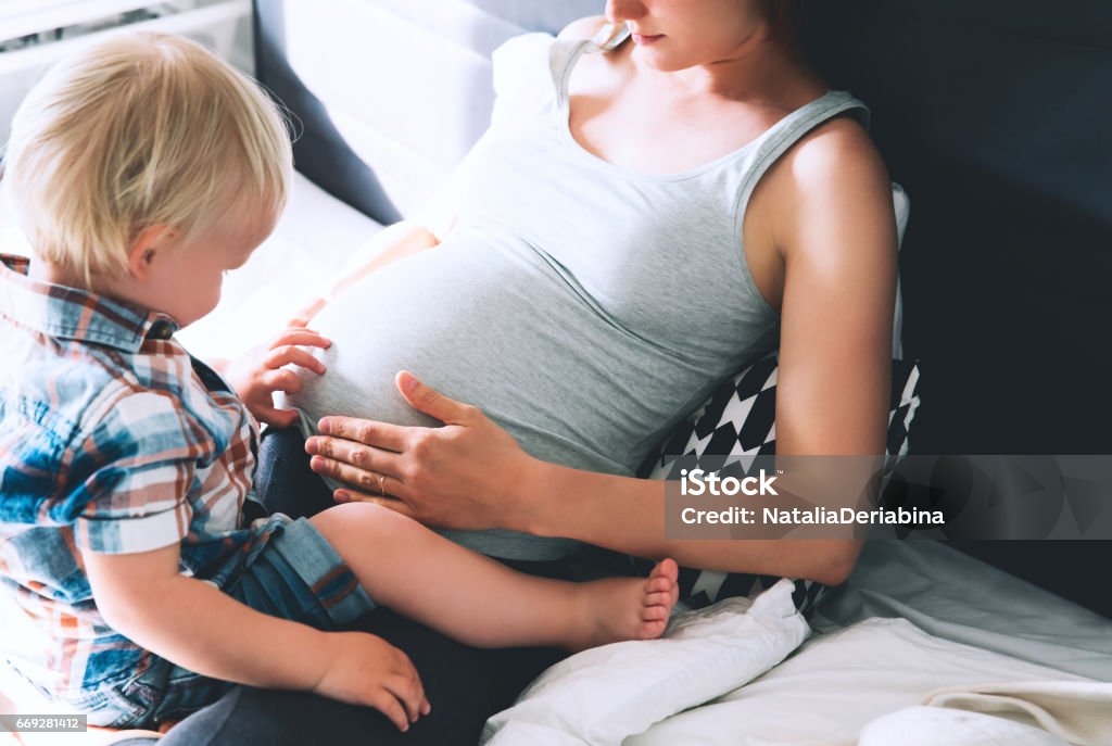 Pregnant mother and son at home. Pregnant mother and son are talking and spending time together in bed at home. Little child boy looking at her mother pregnant tummy. Pregnancy, family, parenthood, preparation and expectation concepts. Pregnant Stock Photo