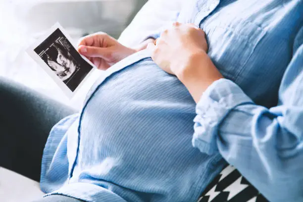 Pregnant woman keeping hand on belly and holding ultrasound image at home interiors. Pregnancy, parenthood, preparation and expectation concept. Close-up, copy space, indoors.