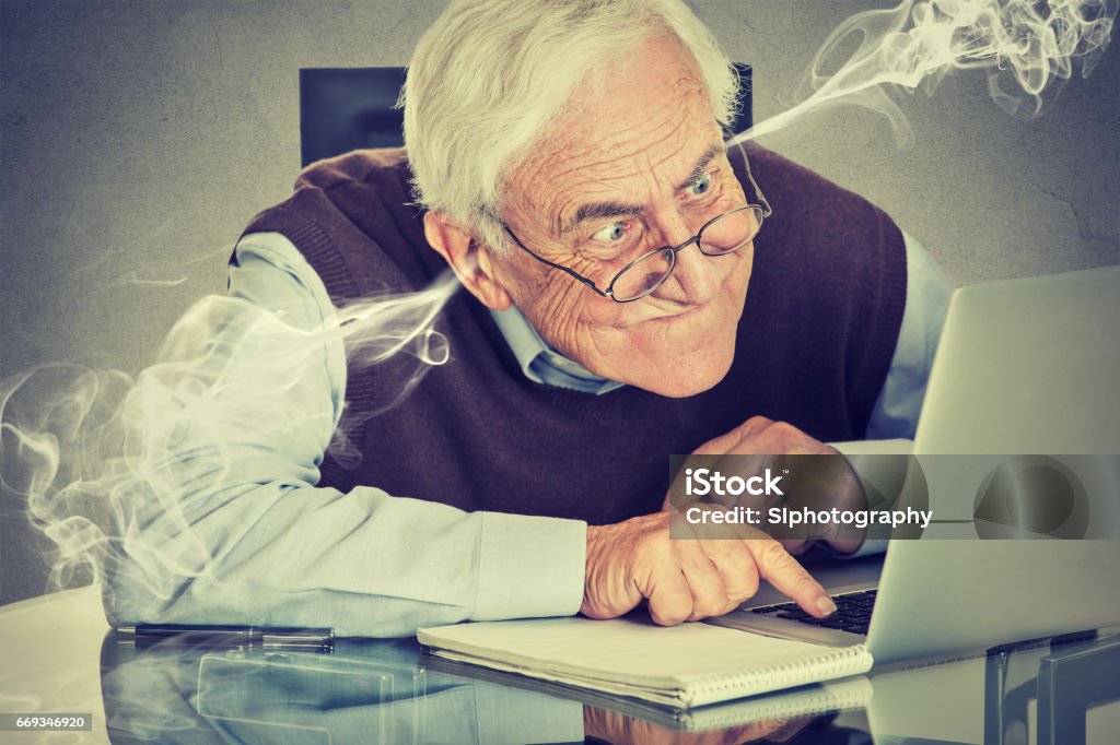 Stressed elderly old man using computer blowing steam from ears Stressed elderly old man using computer blowing steam from ears. Frustrated guy sitting at table working on laptop isolated on gray wall background. Senior people and technology concept Senior Adult Stock Photo