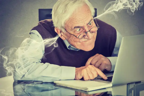 Stressed elderly old man using computer blowing steam from ears. Frustrated guy sitting at table working on laptop isolated on gray wall background. Senior people and technology concept