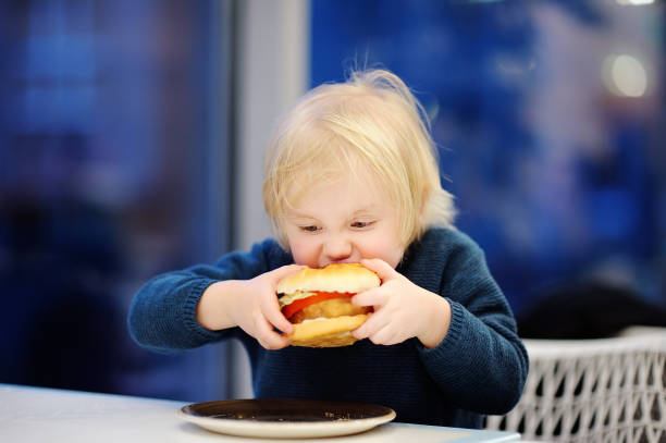 かわいいブロンドの男の子は、ファストフードレストランでハンバーガーを食べます - overweight child eating hamburger ストックフォトと画像