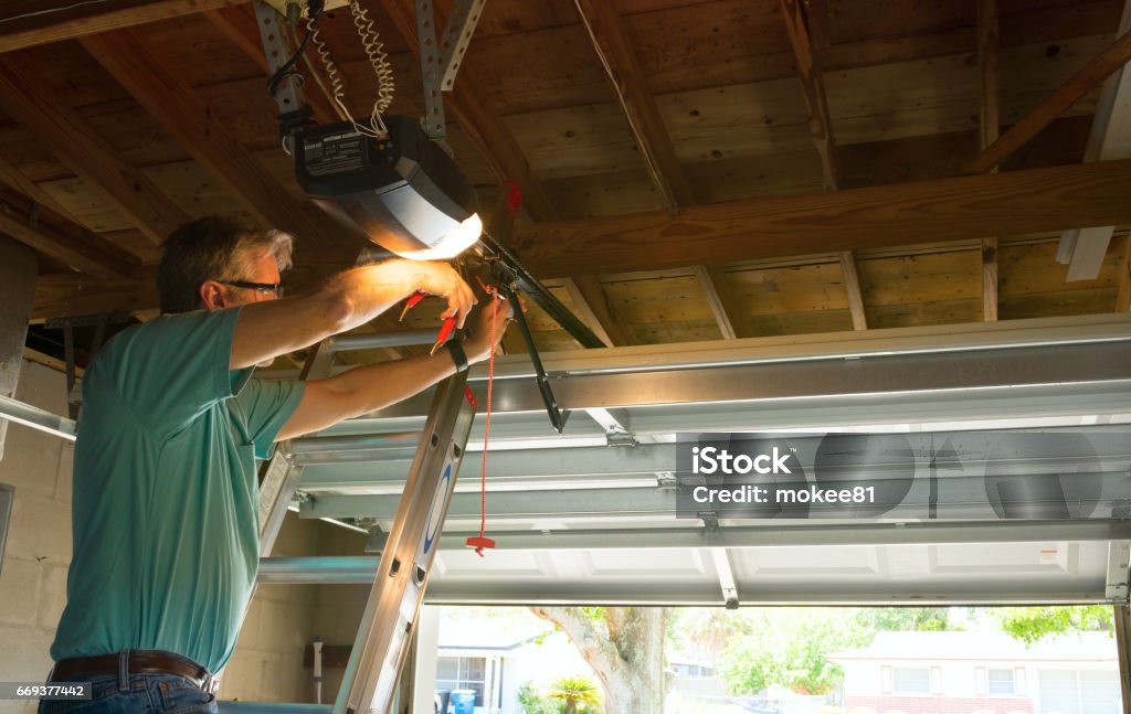 Professional automatic garage door opener repair service technician man working Professional automatic garage door opener repair service technician man working on a ladder at a home residential location making adjustments and fixing it while installing it. Garage Stock Photo