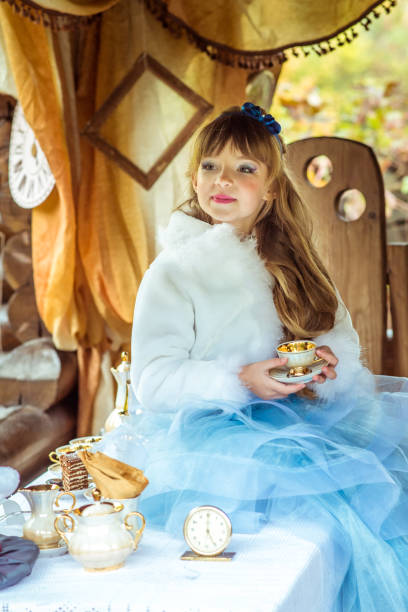 vista frontal de uma menina bonita no cenário da alice no país das maravilhas, segurando uma xícara de chá na mesa - little girls alice in wonderland child fairy tale - fotografias e filmes do acervo